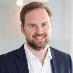 Tim Whitney wearing a white formal shirt, navy blue blazer in front of a blurry background.